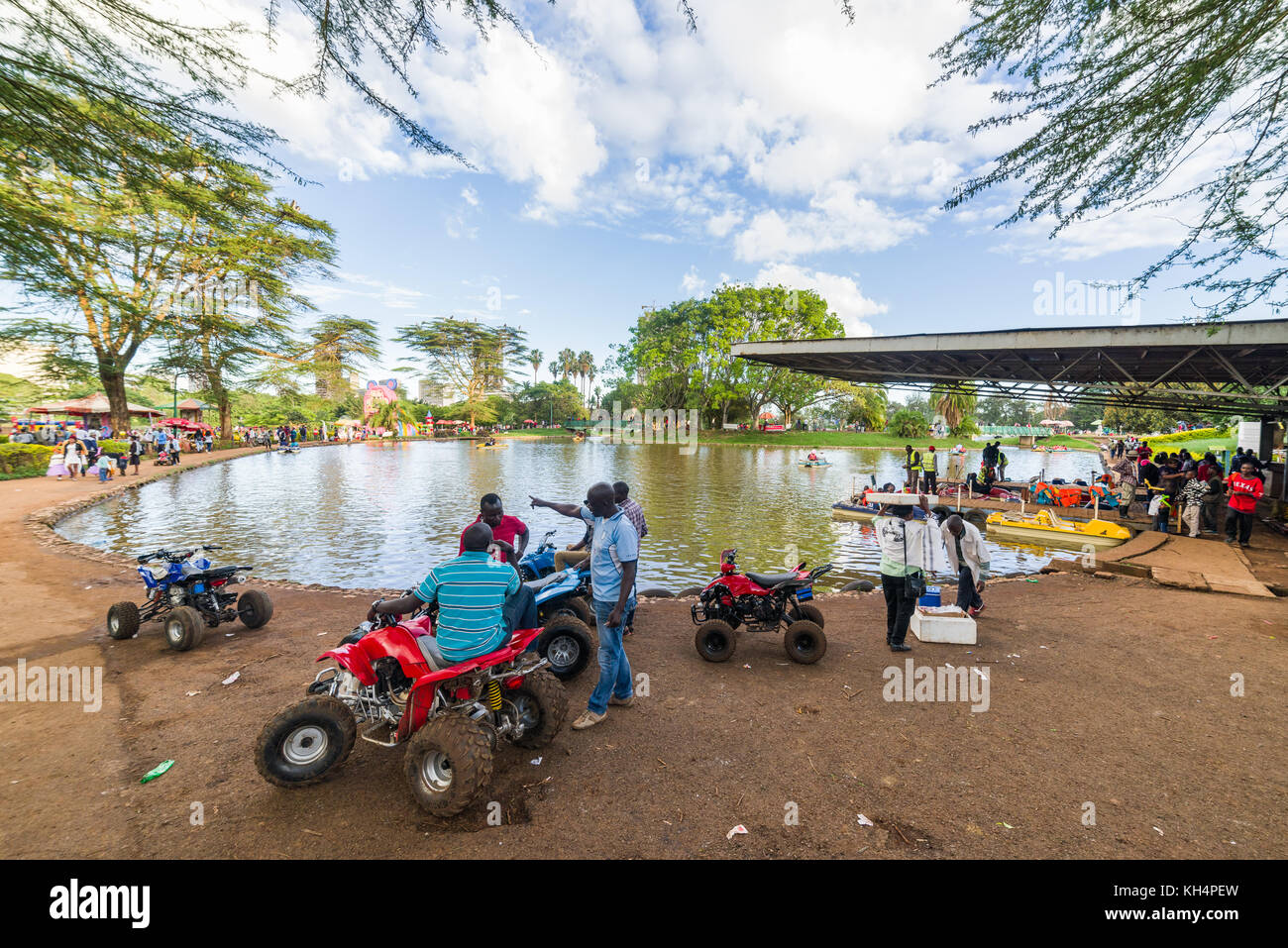 Les gens en premier plan avec des quads pour enfants manèges par lac de plaisance, Uhur Park, Nairobi, Kenya Banque D'Images