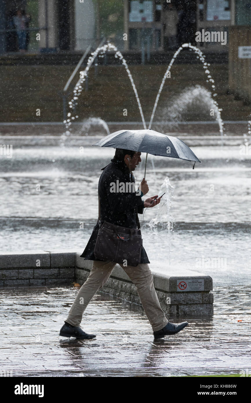 Homme marchant dans les fortes pluies, holding umbrella & utilisation du téléphone, l'adoption de la piscine miroir et fontaines - Bradford City Park, West Yorkshire, Angleterre, Royaume-Uni. Banque D'Images