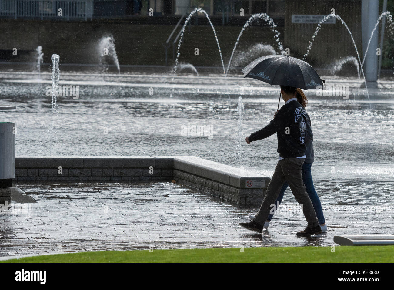 Couple en train de marcher dans de fortes pluies, s'abritant sous un parapluie, l'adoption de la piscine miroir et fontaines - centre de Bradford City Park, West Yorkshire, Angleterre, Royaume-Uni. Banque D'Images