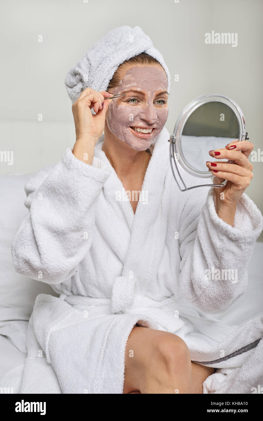 Belle femme portant un masque de beauté tenant un miroir car elle pince à épiler les sourcils Banque D'Images