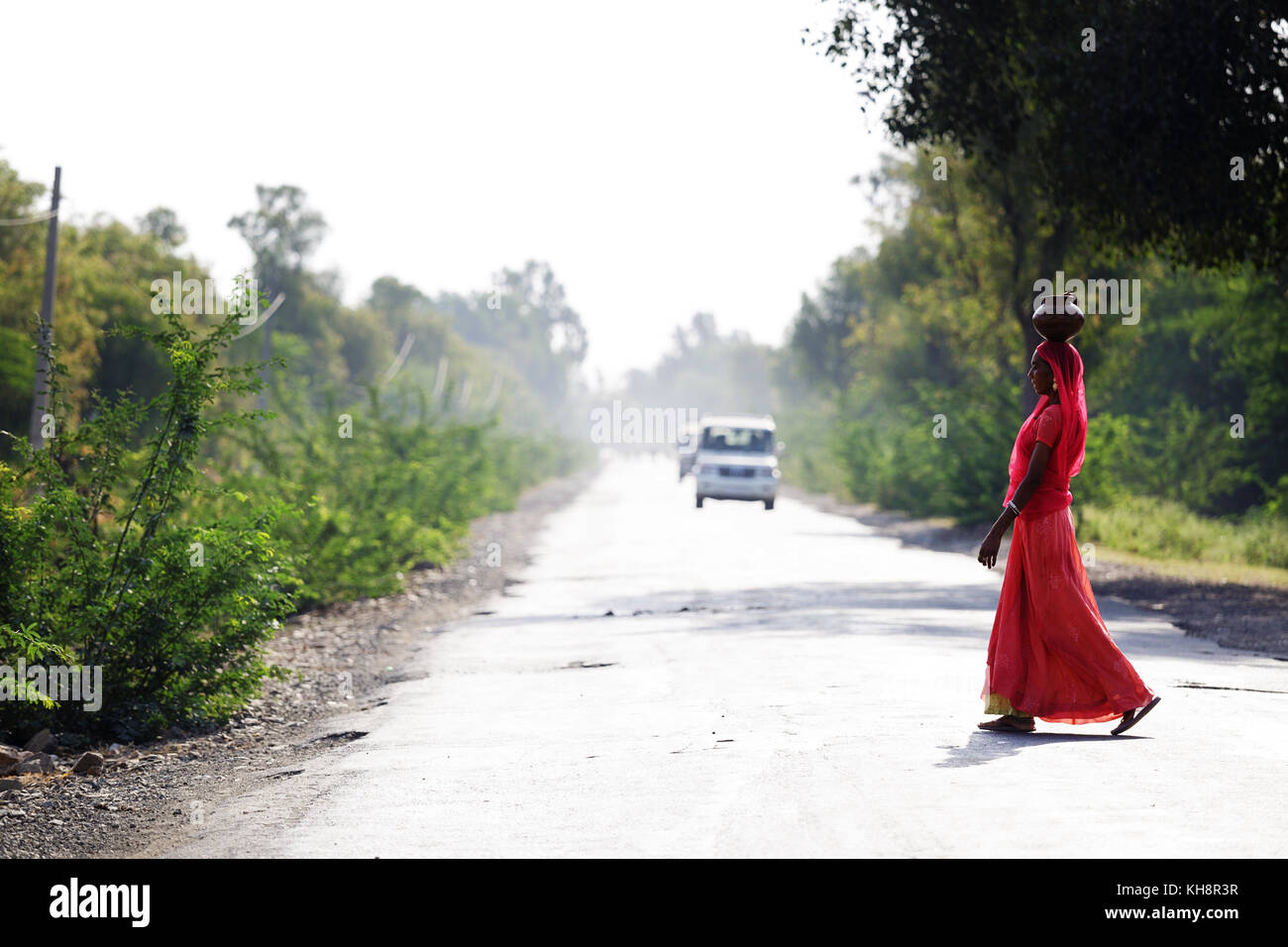 Femme indienne en tenue traditionnelle rouge portant un pot sur sa tête et traverser la route au Rajasthan, Inde. Banque D'Images