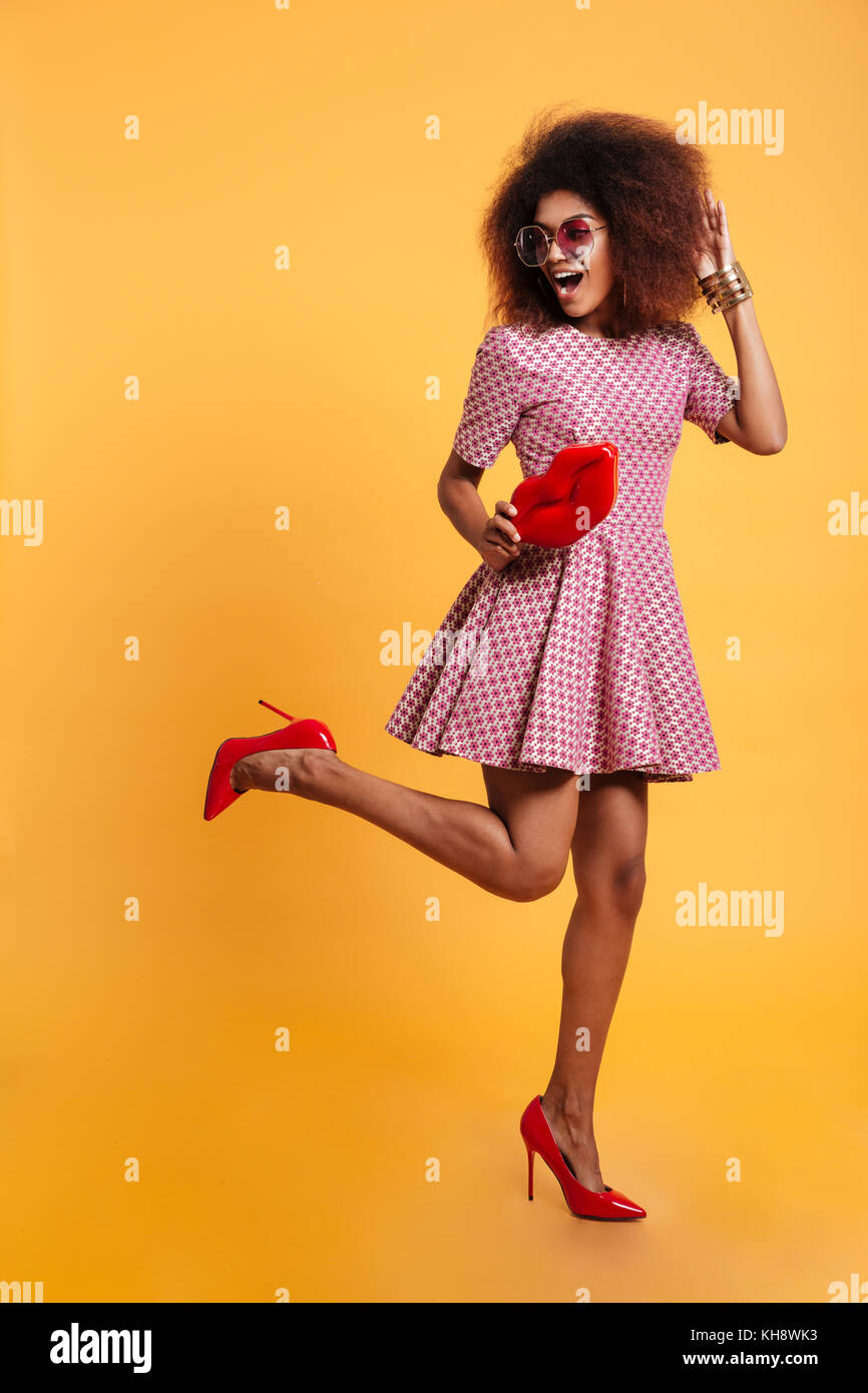 Photo de toute la longueur de l'Afrique de charme élégant rétro femme en robe et talons hauts debout sur une jambe en posant avec big red lips, isolé sur yell Banque D'Images