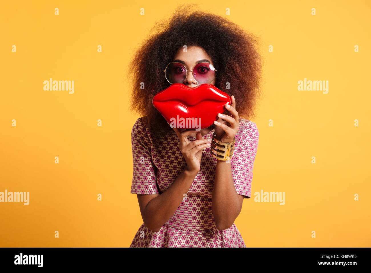 Close-up portrait of African wooman ludique en lunettes holding big red lips devant son visage, looking at camera, isolé sur fond jaune Banque D'Images