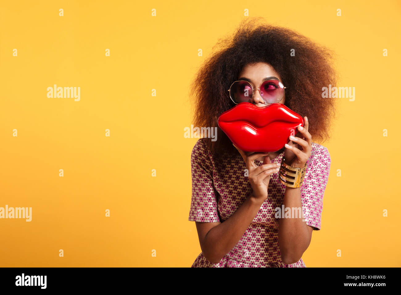 Close-up portrait of african american wooman drôle de lunettes holding big red lips devant son visage, à la recherche de côté, isolé sur fond jaune Banque D'Images