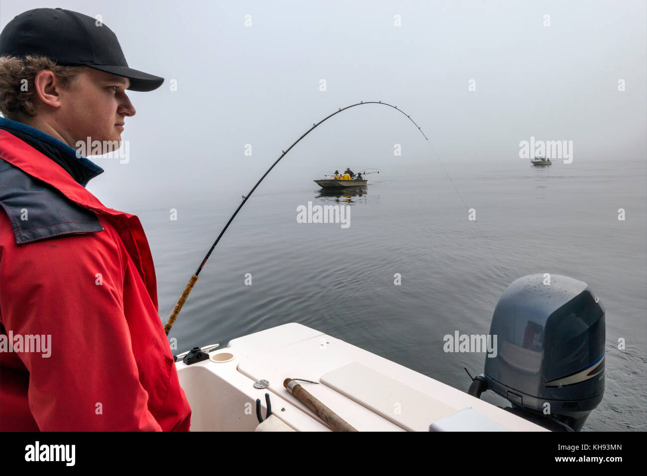 Jeune adulte en veste rouge avec tringle de pêche en bateau, passant d'autres bateaux, matin brouillard dans le détroit de Johnstone au large de l'île de Vancouver, Colombie-Britannique, Can Banque D'Images