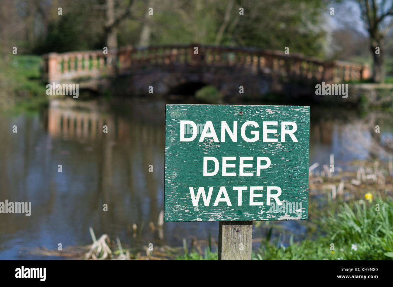 L'eau profonde de danger panneau d'avertissement sur un lac dans le parc du Château Ashby Gardens, hors focus bridge en arrière-plan ; Northamptonshire, Angleterre Banque D'Images