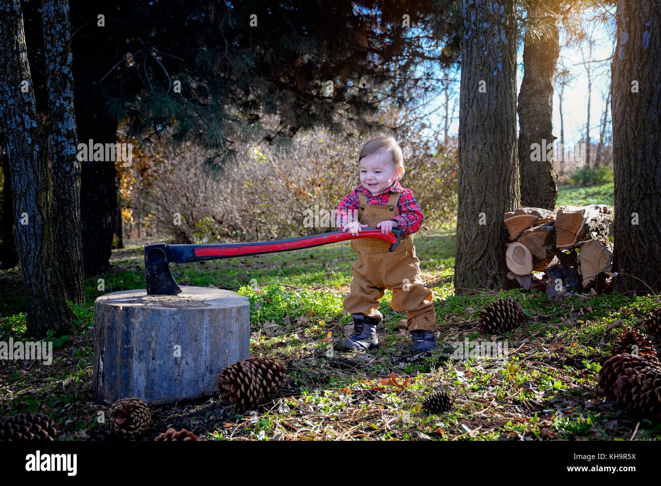 Bébé garçon prétendant être bûcheron porter des bottes et une combinaison à l'aide d'ax prétendant pour couper du bois de cocottes et paramètre de pays rural Banque D'Images