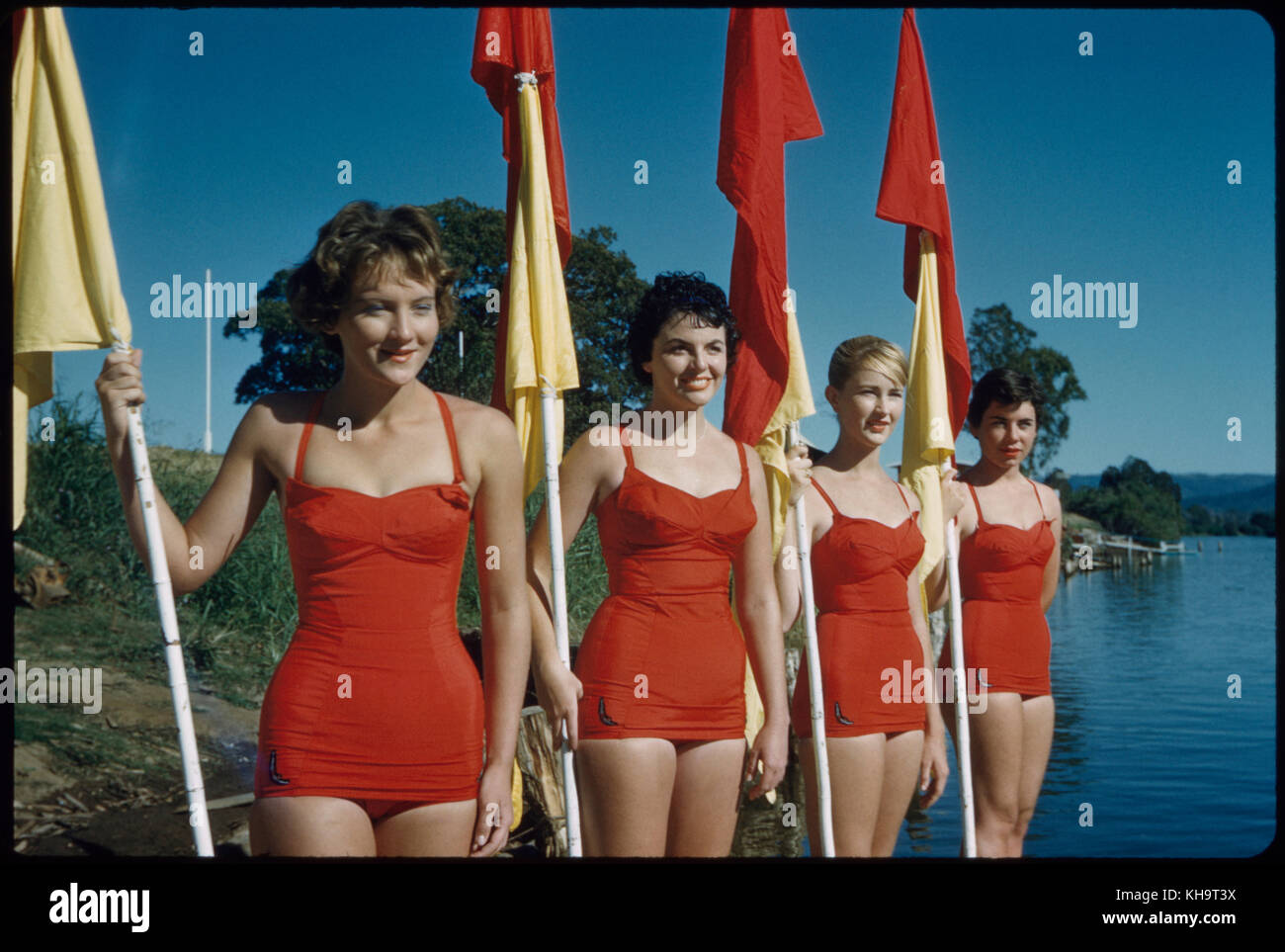 Quatre jeunes femmes adultes portant des maillots de bain rouges d'une pièce et portant des drapeaux rouges et jaunes, Australie, 1960 Banque D'Images
