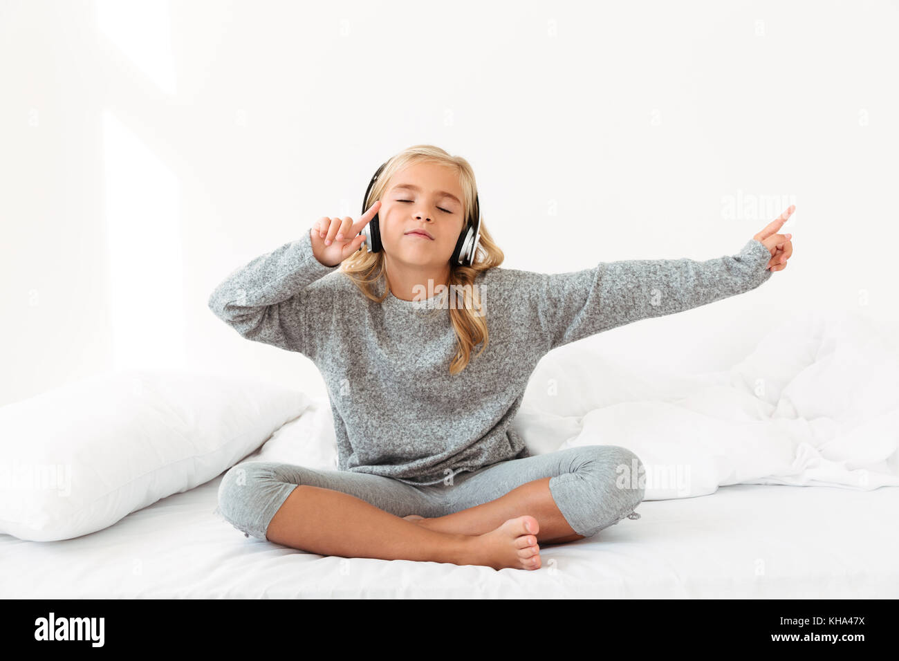 Jolie petite fille en pyjama gris à écouter de la musique avec les yeux fermés, en position assise dans son lit Banque D'Images