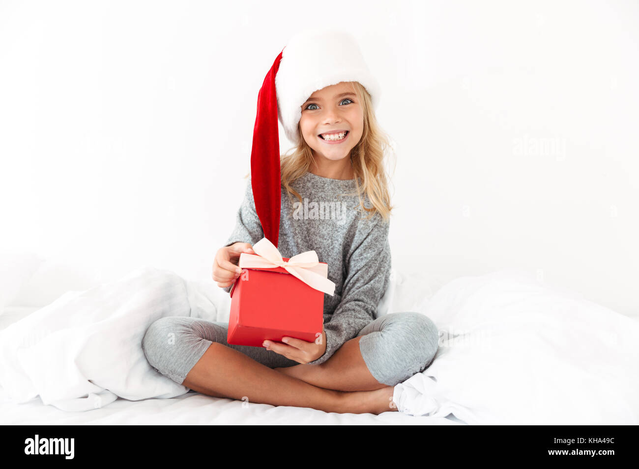 Heureux sort little girl in Santa hat holding gift box, looking at camera while sitting on bed Banque D'Images