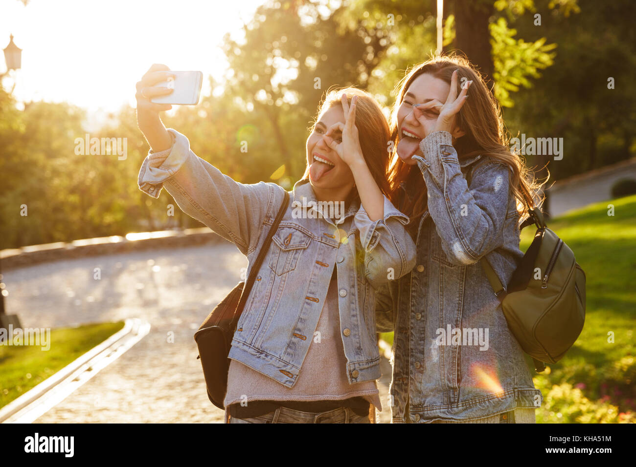 Close-up portrait of two funny friends en regardant à travers l'usure jean décontracté tout en faisant signe OK sur smartphone selfies, extérieur Banque D'Images