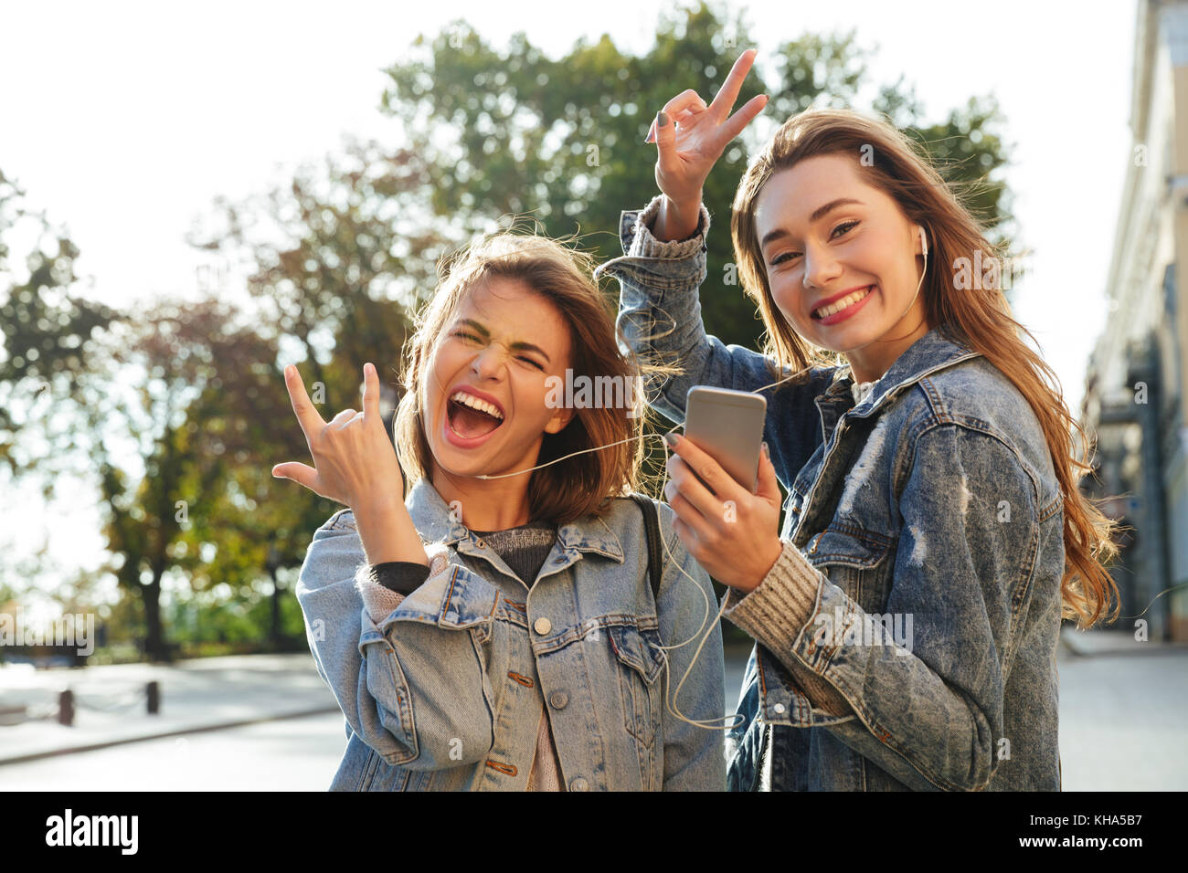 Deux drôles d'amis femme posant tout en écoutant de la musique sur smartphone, extérieur Banque D'Images