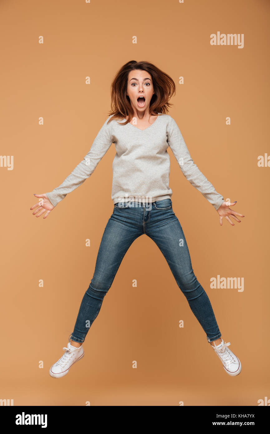 Photo de pleine longueur choqué jeune femme en blouse grise et un jean saute au-dessus de fond beige Banque D'Images