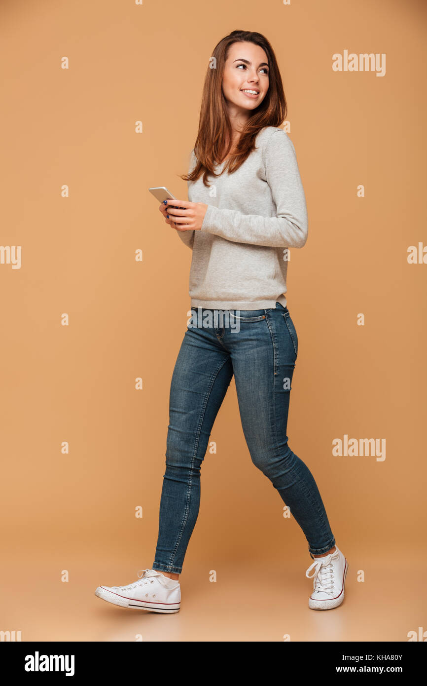 Full Length portrait of smiling brunette woman en blouse grise et un jean, holding smartphone, à côté, isolé sur fond beige Banque D'Images