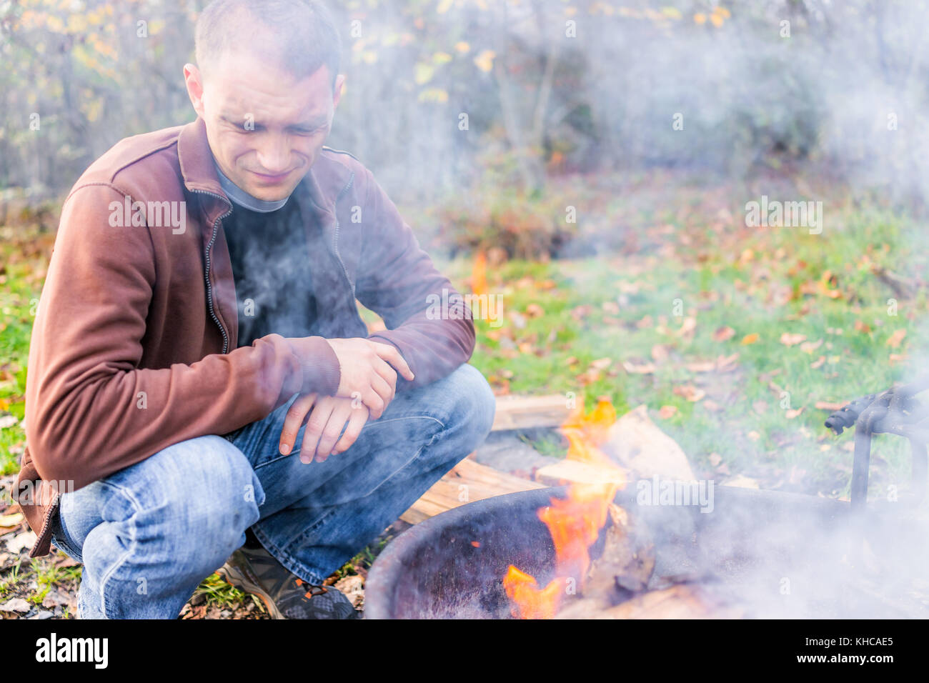 Sale jeune homme réaliste avec visage couvert de cendres d'un feu de camp de soufflage gris en flamme feu luxe par sciage et forêt d'automne, beaucoup de fumée Banque D'Images