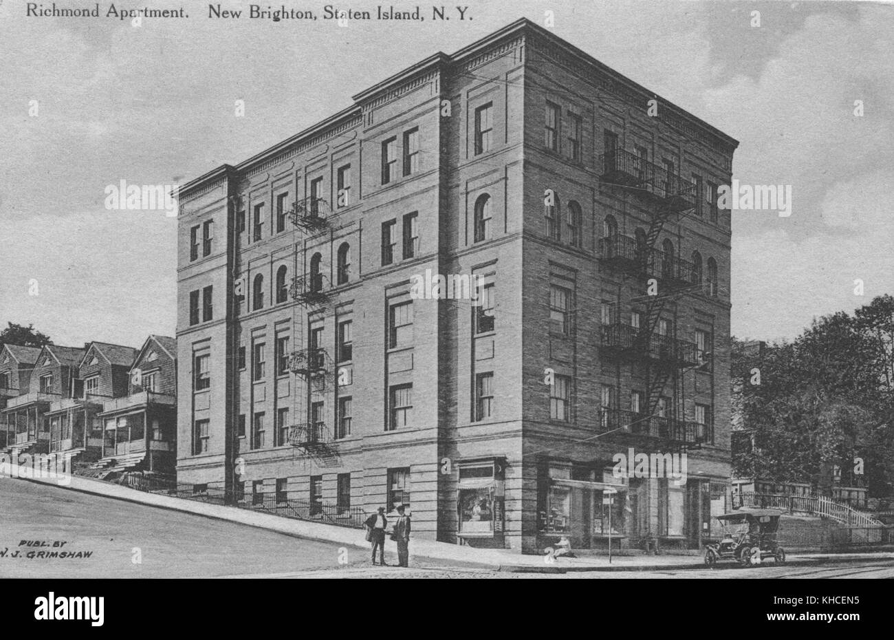 Carte postale d'une photo de la vue extérieure d'un immeuble de cinq étages, deux hommes sont vus conversant dans la rue tandis qu'une voiture ancienne est garée à partir de l'immeuble, Le bâtiment lui-même est une structure en pierre située au bas d'une petite colline où d'autres maisons peuvent être vues le long de la rue, New Brighton, Staten Island, New York, 1900. De la bibliothèque publique de New York. Banque D'Images