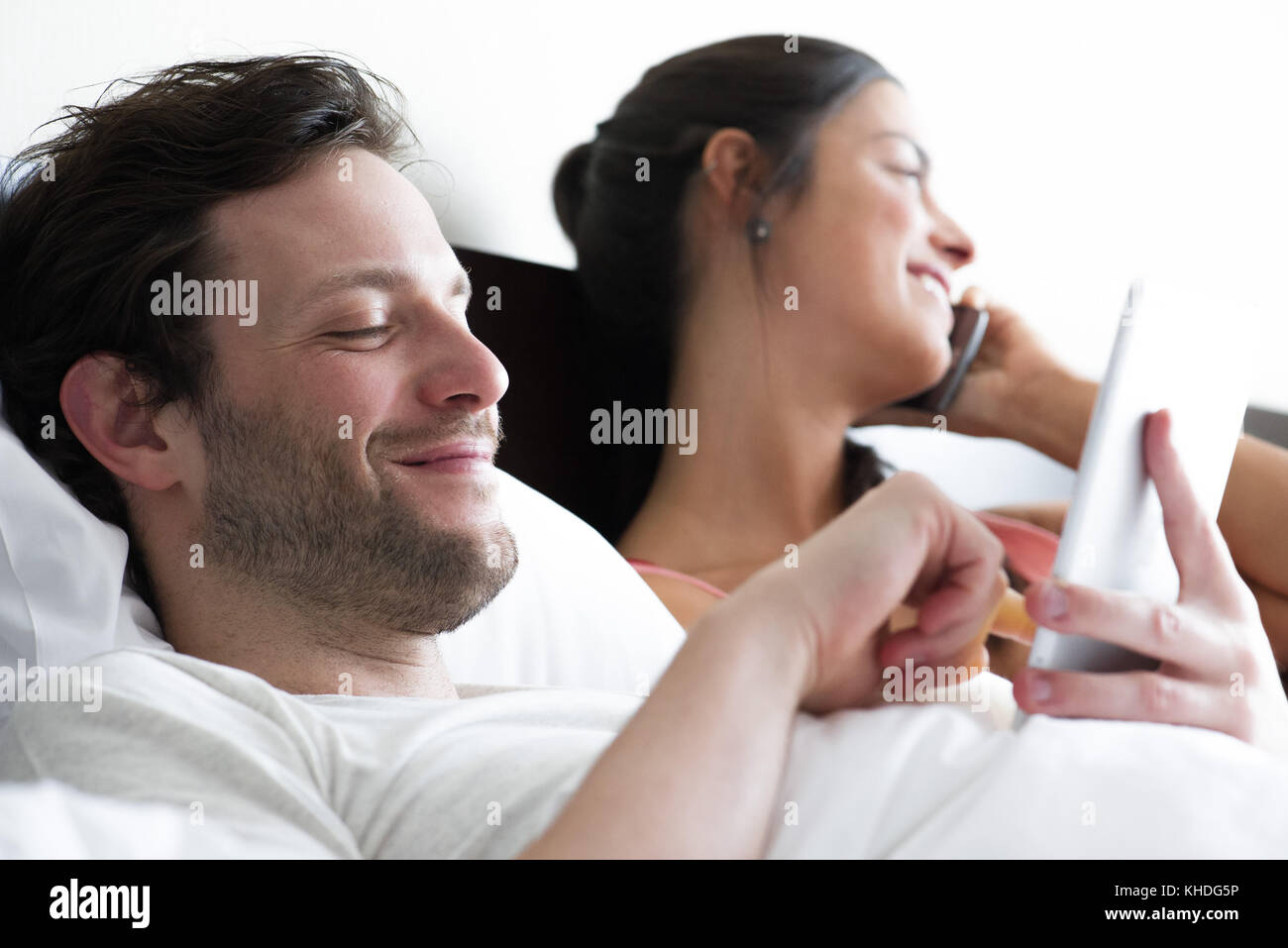 Couple relaxing in bed, man using digital tablet while femme fait appel Banque D'Images
