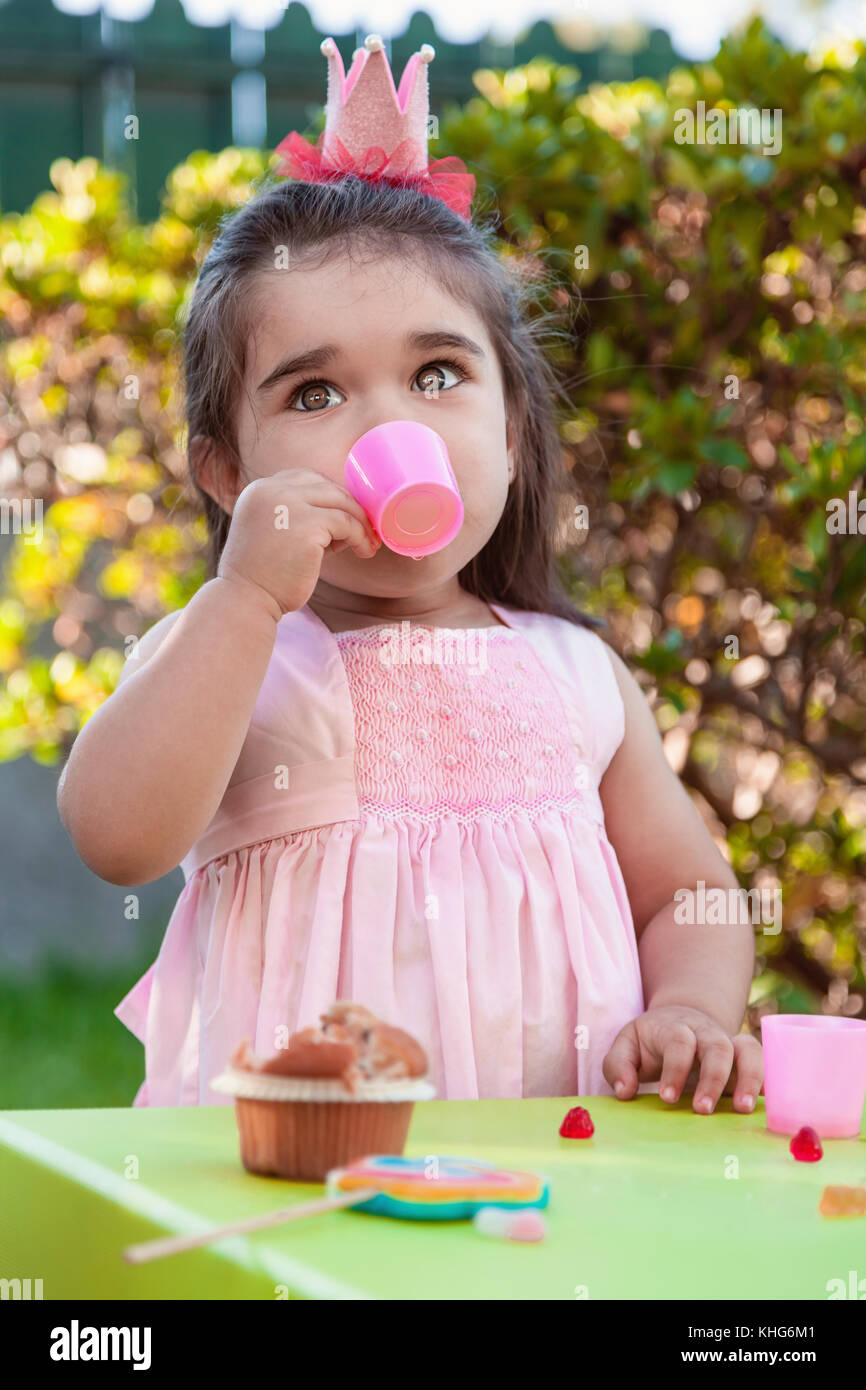 Baby girl playing tea party en plein air dans l'eau de tasse avec Lucette, muffin et gummies sur table. robe rose et la reine ou princesse couronne Banque D'Images