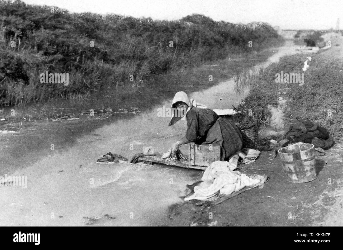 Une photographie d'une femme faisant la blanchisserie dans un petit plan d'eau courante, elle porte des vêtements simples et un chapeau alors qu'à genoux dans la boue au bord de l'eau, l'herbe pousse court le long du bord de l'eau, Camargue, France, juillet, 1922. Banque D'Images