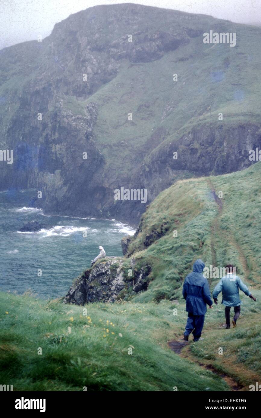 Une photo de deux Randonneurs marchant le long d'une baignoire étroite en haut d'une falaise couverte d'herbe, une autre personne peut être vu assis sur un affleurement rocheux de l'avant des deux autres randonneurs, les vagues de l'océan peut être vu s'écraser contre le bas de la falaise, 1965. Banque D'Images