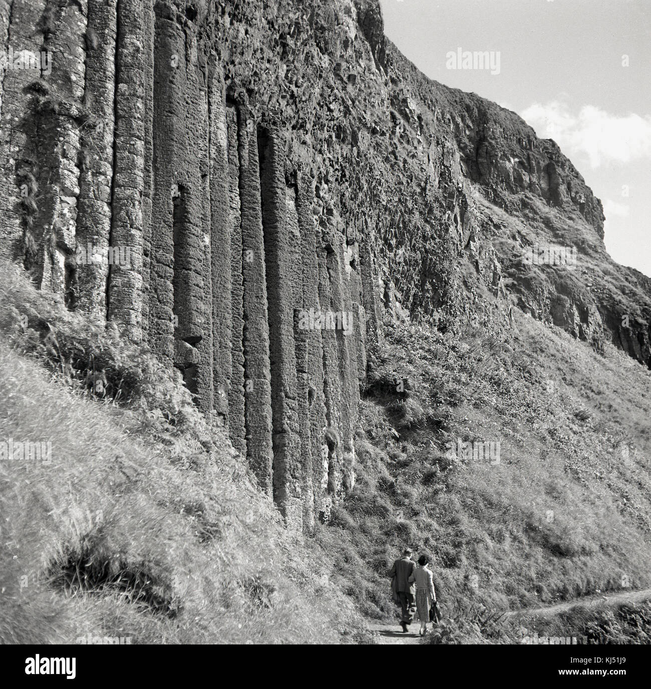 Années 1950, historique, un couple marcher ensemble le long d'un étroit sentier littoral à côté d'une falaise abrupte montrant les anciennes colonnes de basalte volcanique et de formations rocheuses qui font de la Giant's Causeway en Co Antrim, en Irlande du Nord, un endroit si spécial à visiter. Banque D'Images