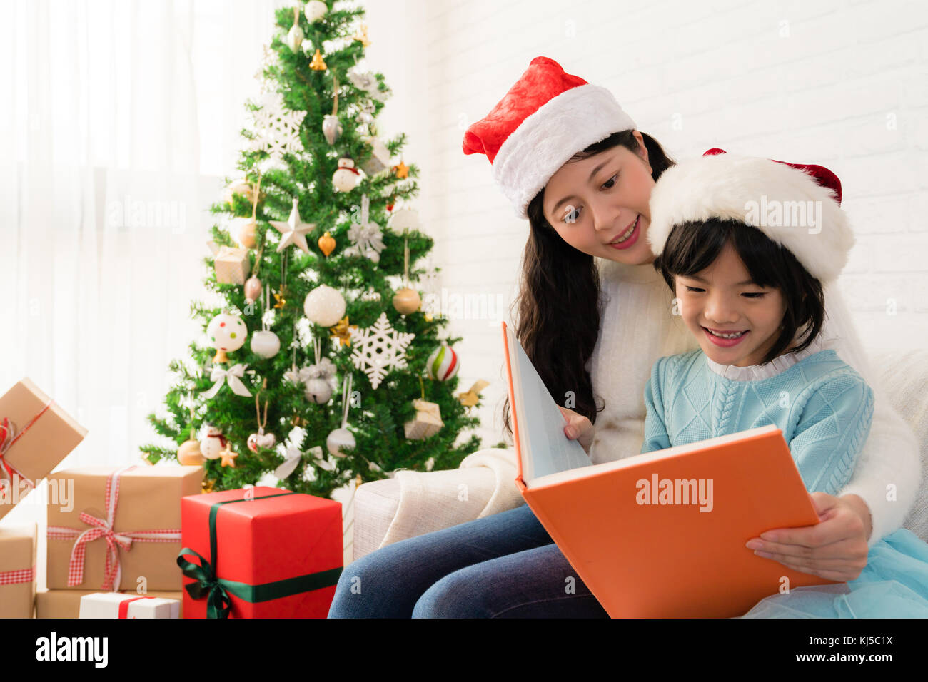 Jeune maman asiatique lire un livre à son joli fille près de l'arbre de Noël à l'intérieur à la maison. Joyeux Noël et de bonnes vacances ! Banque D'Images