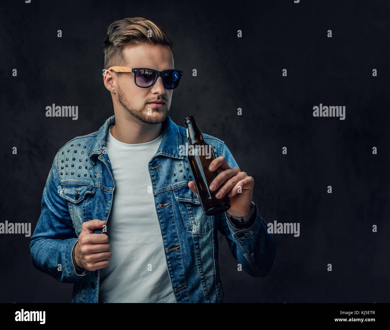 Homme blond positif vêtu d'une veste en jean et des lunettes est titulaire d'une bouteille avec de la bière artisanale. Banque D'Images