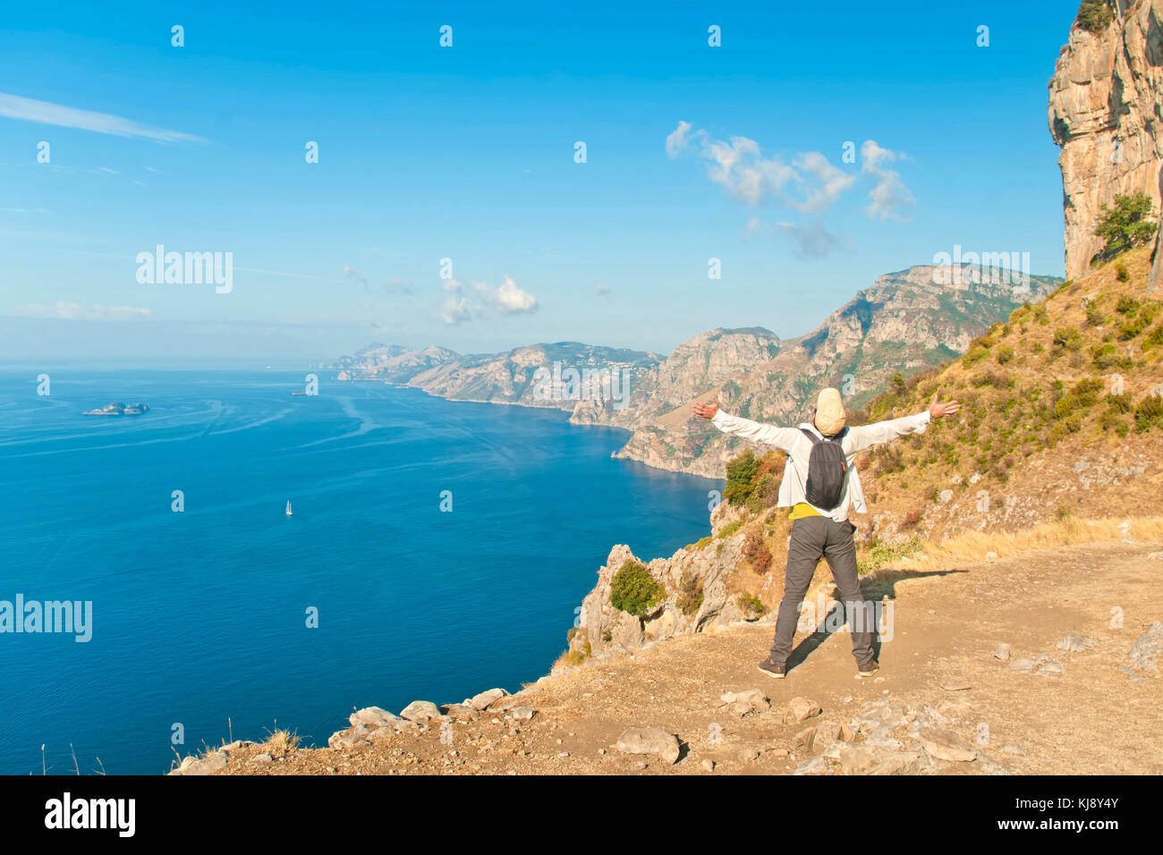 Jeune homme en chemise blanche et touristique cap debout à bord de la falaise avec ses bras ouverts à la recherche en mer et montagnes sur le chemin des dieux, côte amalfitaine Banque D'Images