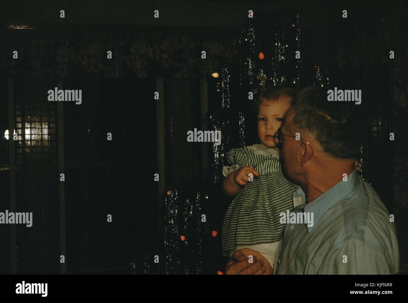 Une photographie d'un grand-père tenant son enfant en petite-fille, elle porte une robe rayée verte et blanche, tandis qu'il porte une chemise bleu enfoncé, guirlandes et lumières peuvent être vu suspendu à l'arbre de Noël derrière eux, 1952. Banque D'Images