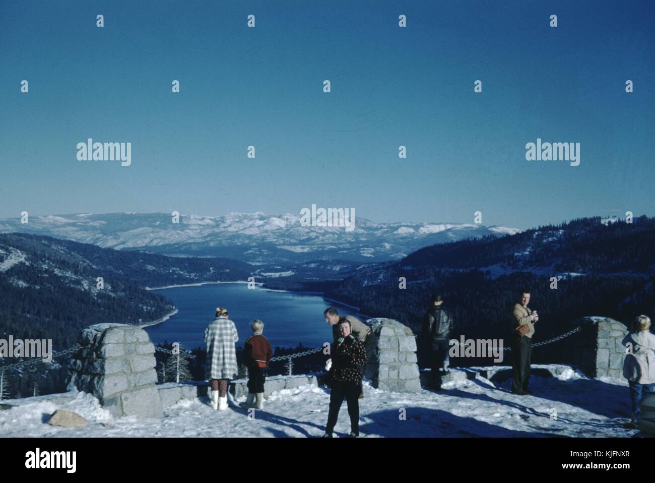 Une photographie d'hommes, de femmes et d'enfants dans une zone d'observation, il y a de la neige qui couvre le sol et tout le monde porte des vêtements d'hiver, la zone d'observation surplombe un grand lac bleu et des montagnes couvertes d'arbres et de neige, 1952. Banque D'Images