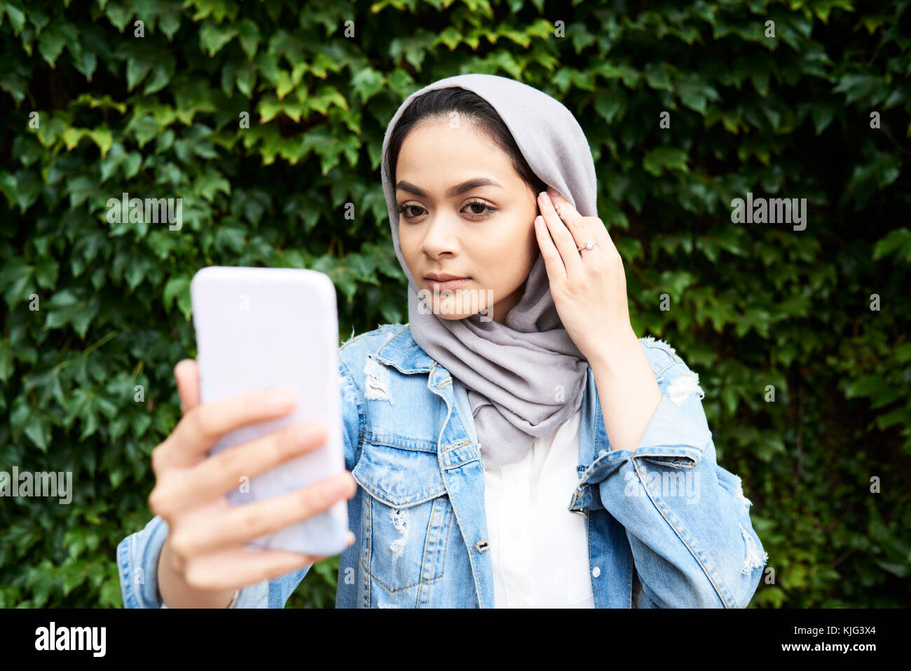 Londres, Royaume-Uni, Angleterre, belle jeune fille musulmane à son téléphone à l'écran portant le hijab contre mur de verdure, étudiant de l'université musulmane Banque D'Images