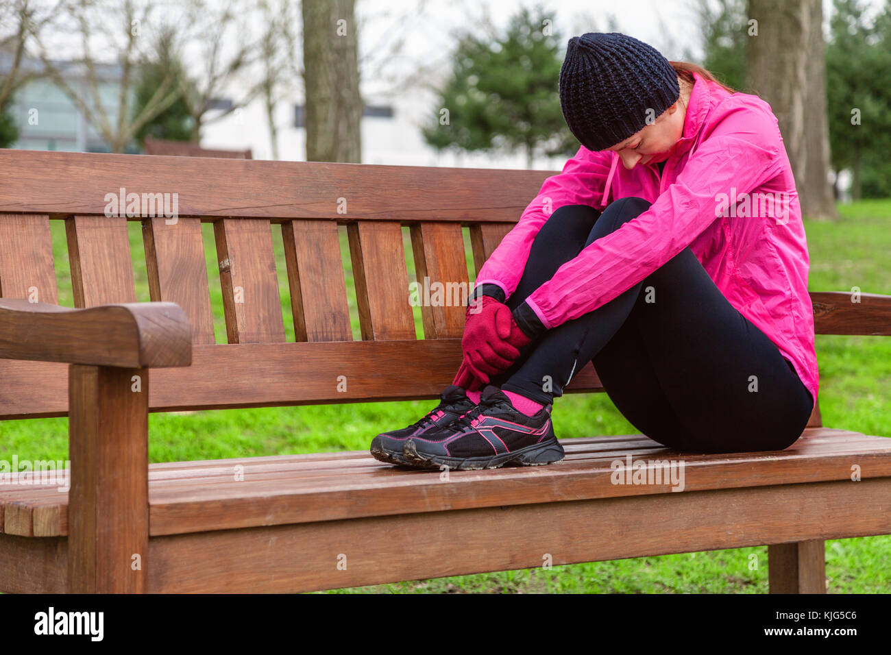 Femme athlète fatigué ou déprimé se reposant sur un banc sur une froide journée d'hiver sur la voie de la formation d'un parc urbain. Jeune femme portant un coupe-vent rose Banque D'Images