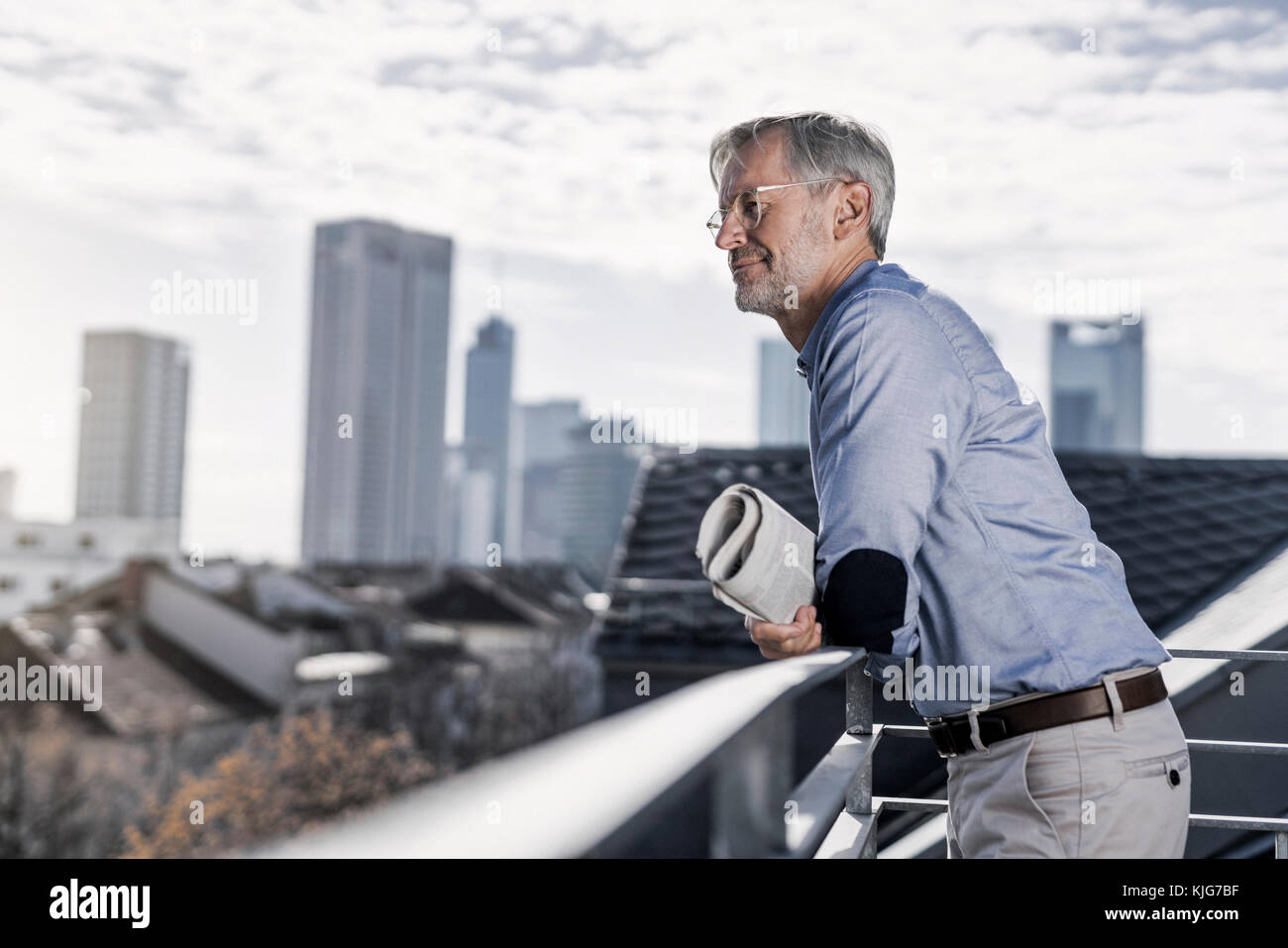 Homme aux cheveux gris se tenant debout sur un balcon avec vue sur la ville à la recherche de journaux Banque D'Images Homme aux cheveux gris se tenant debout sur un balcon avec vue sur la ville à la recherche de journaux Banque D'Images