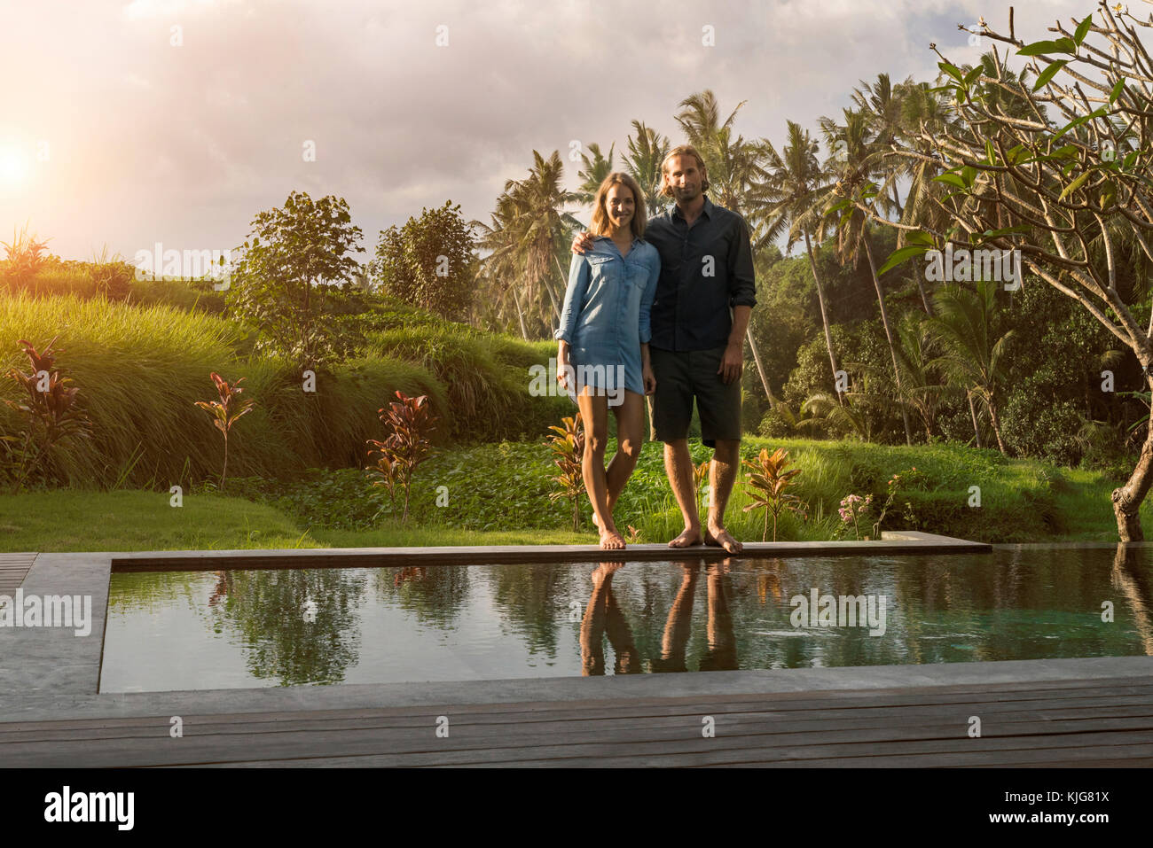 Smiling couple debout sur le bord d'une piscine dans un jardin tropical luxuriant Banque D'Images Smiling couple debout sur le bord d'une piscine dans un jardin tropical luxuriant Banque D'Images
