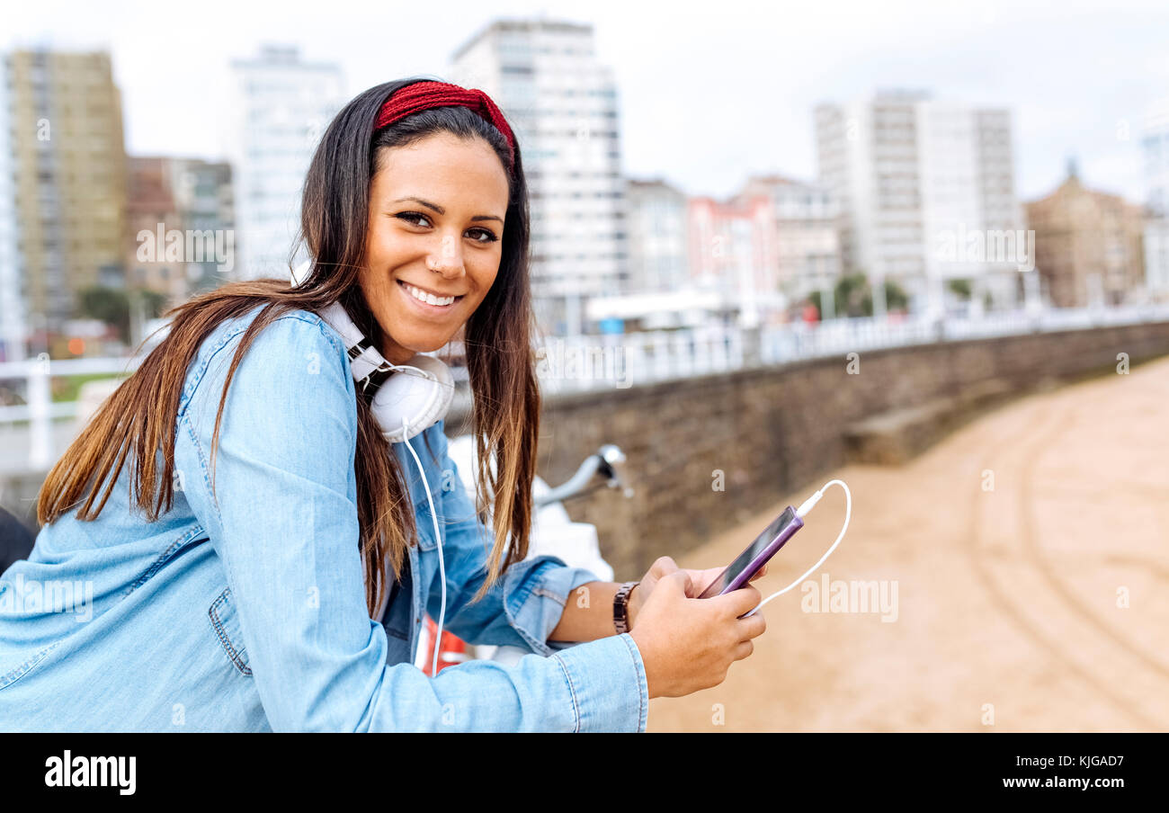 L'Espagne, Gijon, smiling young woman with cell phone et de casques à promenade au bord de l'eau Banque D'Images
