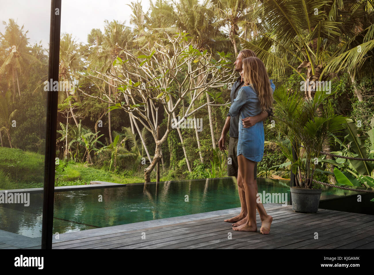 Hugging couple standing sur terrasse en bois en face de la piscine et bénéficiant de superbes vue dans jardin tropical luxuriant Banque D'Images Hugging couple standing sur terrasse en bois en face de la piscine et bénéficiant de superbes vue dans jardin tropical luxuriant Banque D'Images