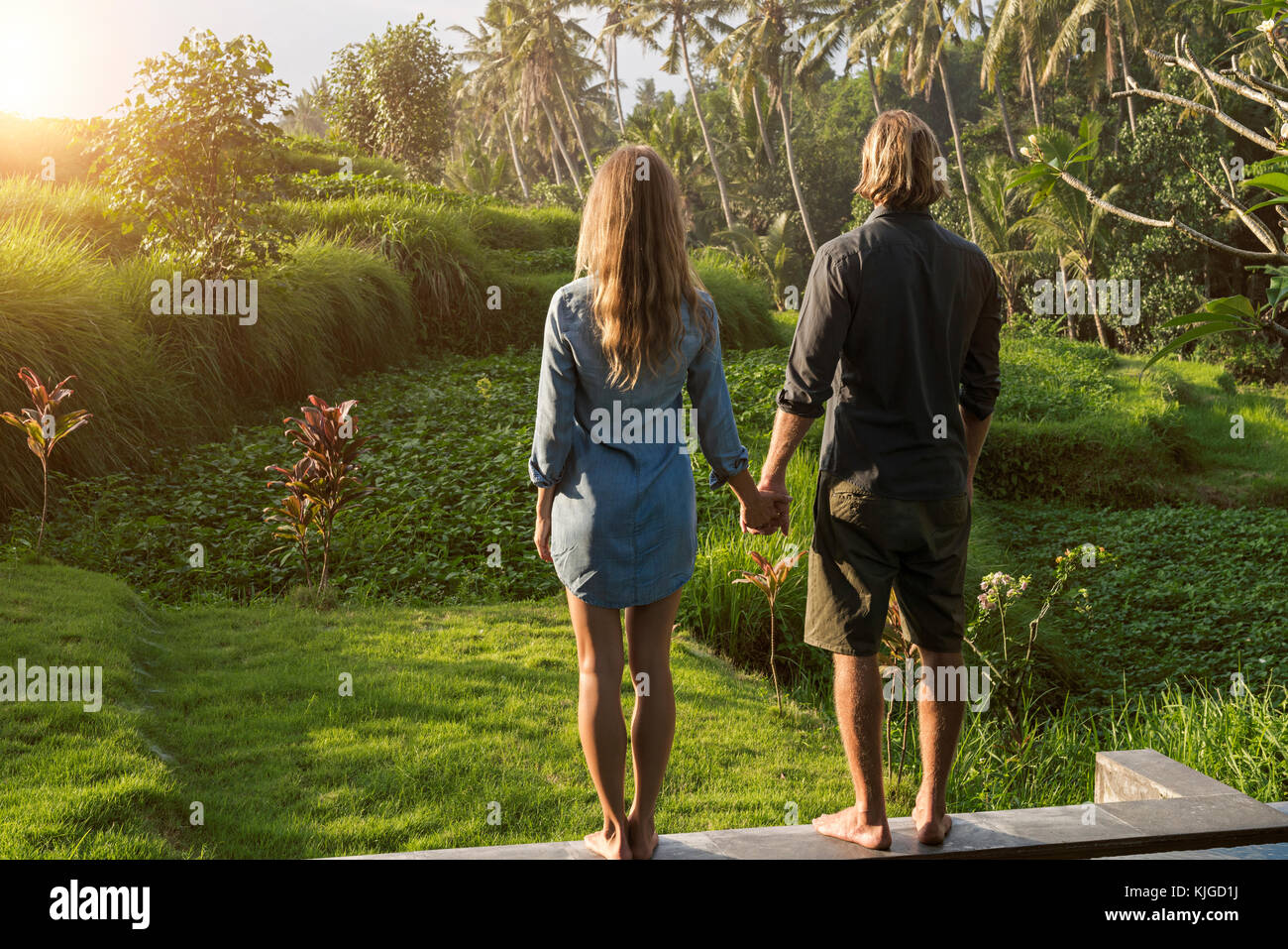 Couple holding hands en étant debout sur le bord d'une piscine et bénéficiant d'une vue imprenable sur le coucher du soleil dans le jardin tropical luxuriant Banque D'Images Couple holding hands en étant debout sur le bord d'une piscine et bénéficiant d'une vue imprenable sur le coucher du soleil dans le jardin tropical luxuriant Banque D'Images