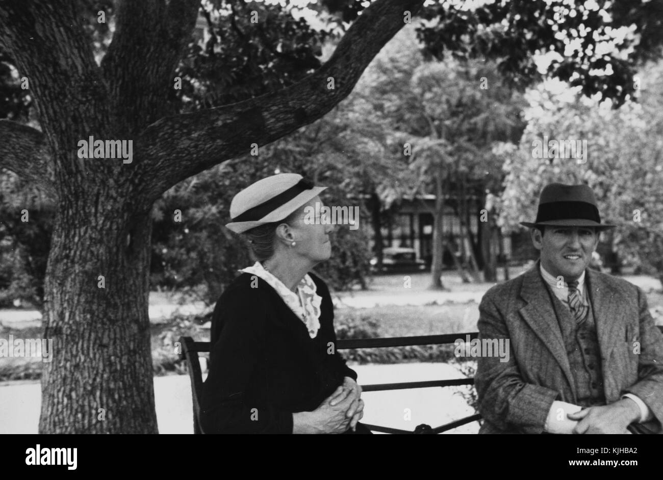 Une photographie d'une femme et un homme assis sur un banc de parc en métal, la femme porte une robe sombre et un chapeau, l'homme porte un costume gris et une cravate avec un chapeau, des arbres, l'herbe, et des chemins de randonnée par le biais de Jackson Square peut être vu derrière eux, New Orleans, Louisiane, 1935. à partir de la bibliothèque publique de new york. Banque D'Images