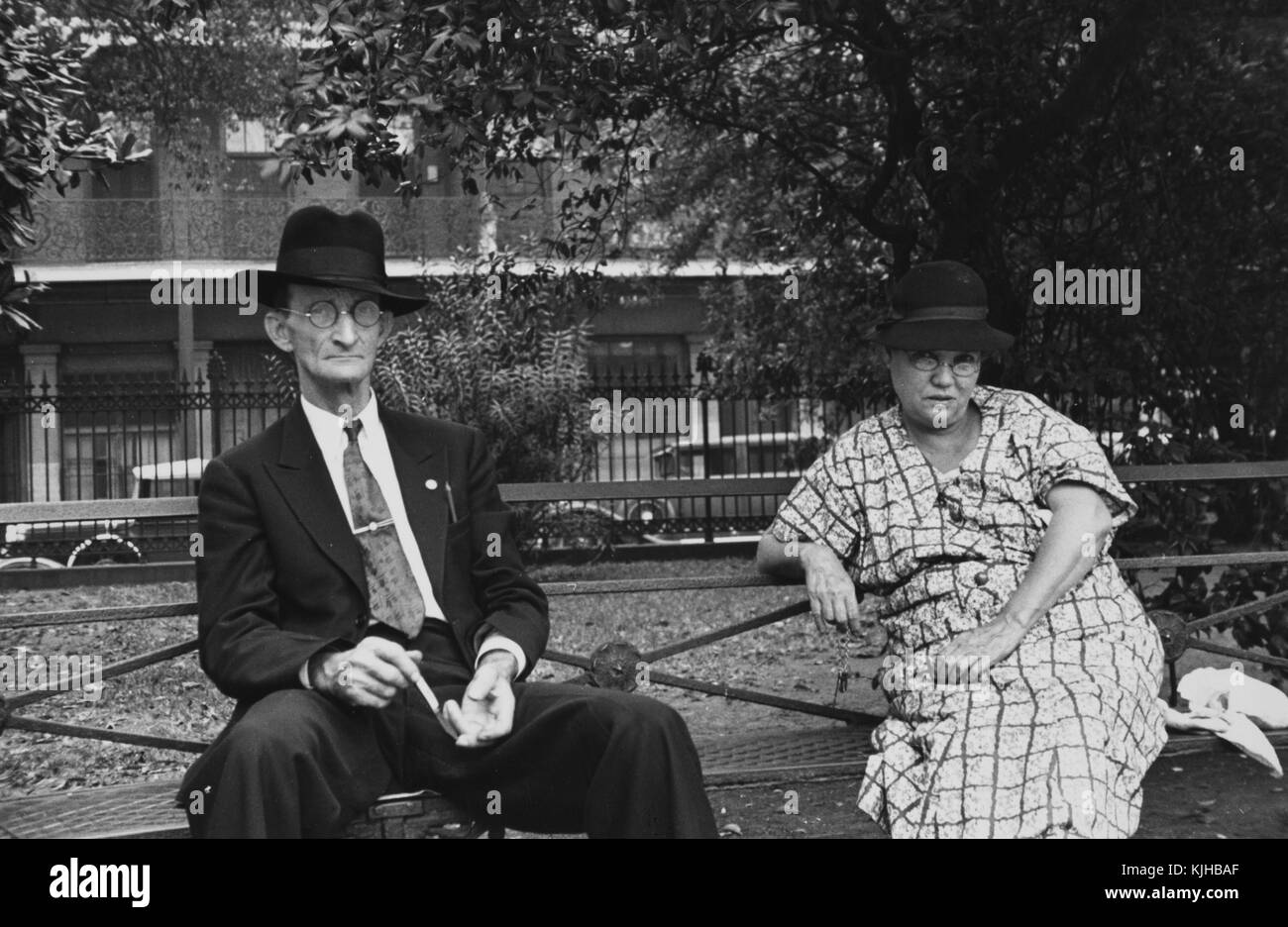 Un homme et une femme assis sur un banc de parc en métal, l'homme porte un  costume et une cravate avec un chapeau, la femme porte une robe à motifs et  un