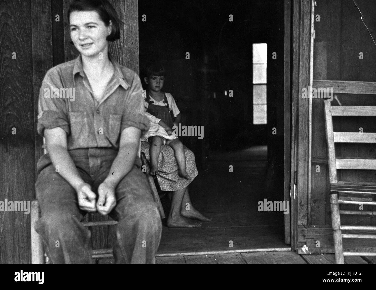 Photographie en noir et blanc d'une famille de l'époque de la dépression métayer, femme assise sur une chaise, au premier plan, une seconde femme tenant deux petits enfants, derrière elle, à l'intérieur d'un bâtiment, de l'Arkansas, 1935. à partir de la bibliothèque publique de new york. Banque D'Images
