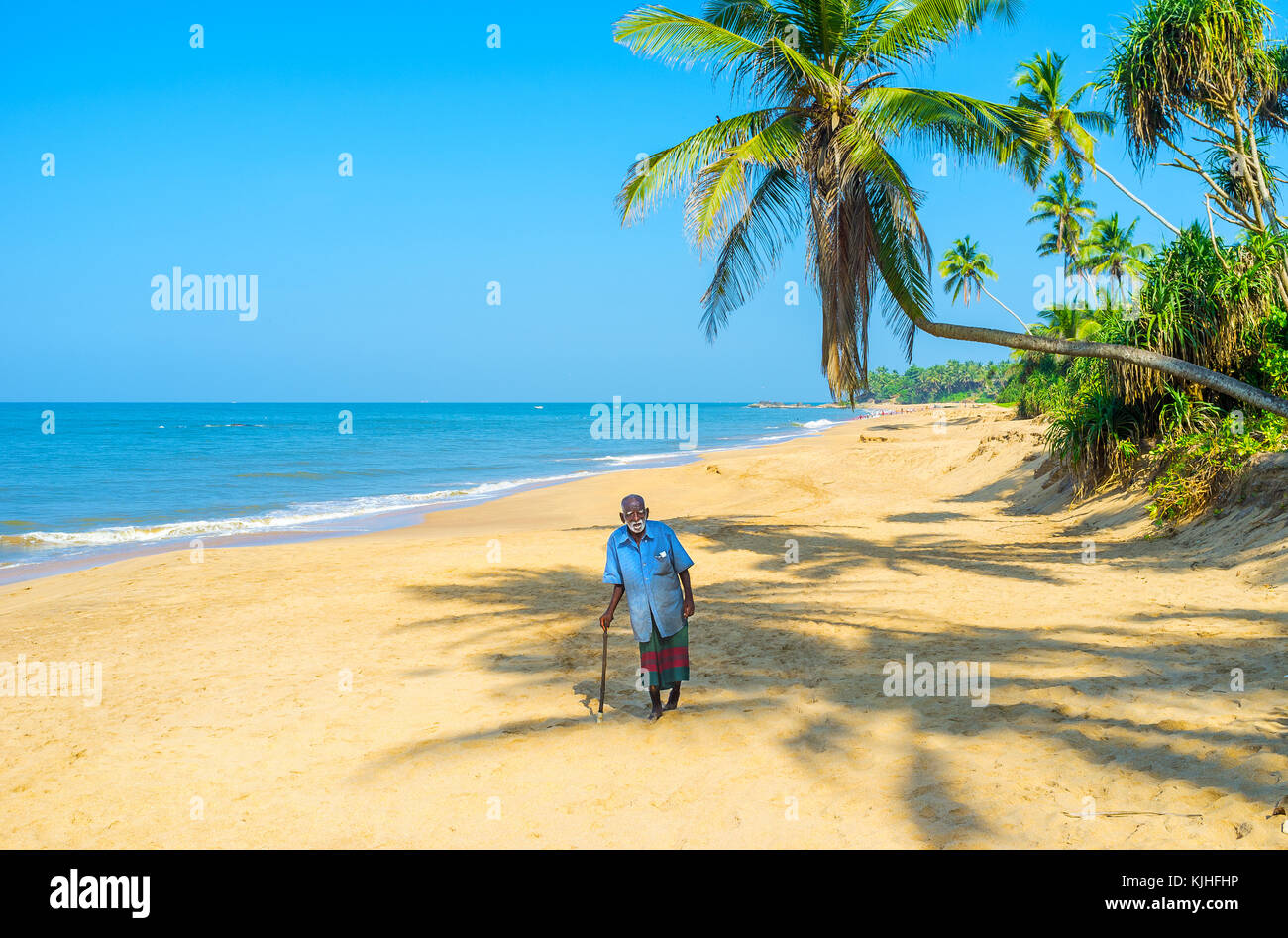 Bentota, Sri Lanka - 6 décembre 2016 : le vieux pêcheur à pied avec un bâton le long de la plage paradisiaque de palmiers et des eaux bleues de l'Inde Banque D'Images