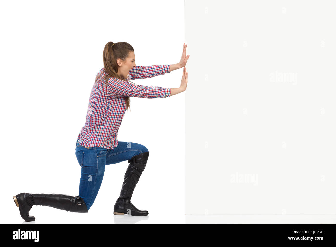 Jeune femme en jeans, bottes noires et chemise de bûcheron poussant un mur blanc et criant. Vue de côté, pleine longueur portrait isolé sur blanc. Banque D'Images