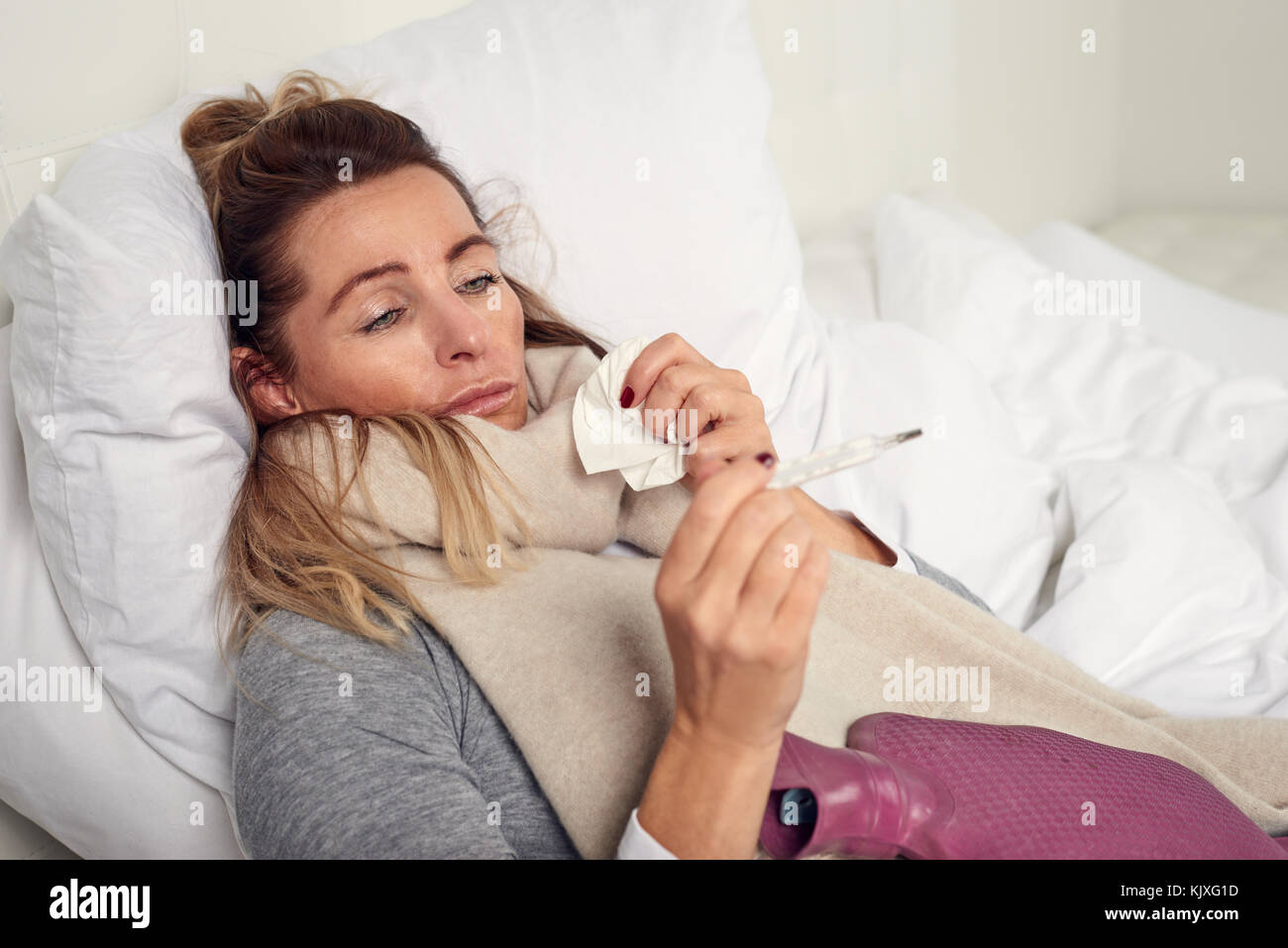 Femme malade avec une expression misérable de prendre sa température pendant qu'elle se trouve au lit souffrant d'un rhume et de la grippe saisonnière en essayant de garder au chaud Banque D'Images
