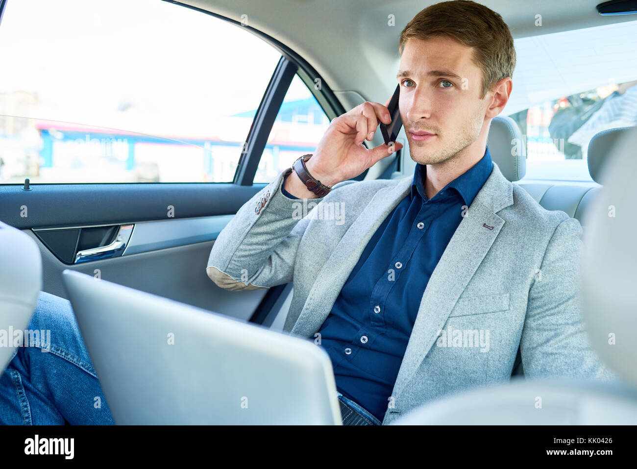 Portrait de jeune homme parlant par téléphone et à l'aide d'ordinateur portable assis sur siège arrière de voiture de luxe, copy space Banque D'Images