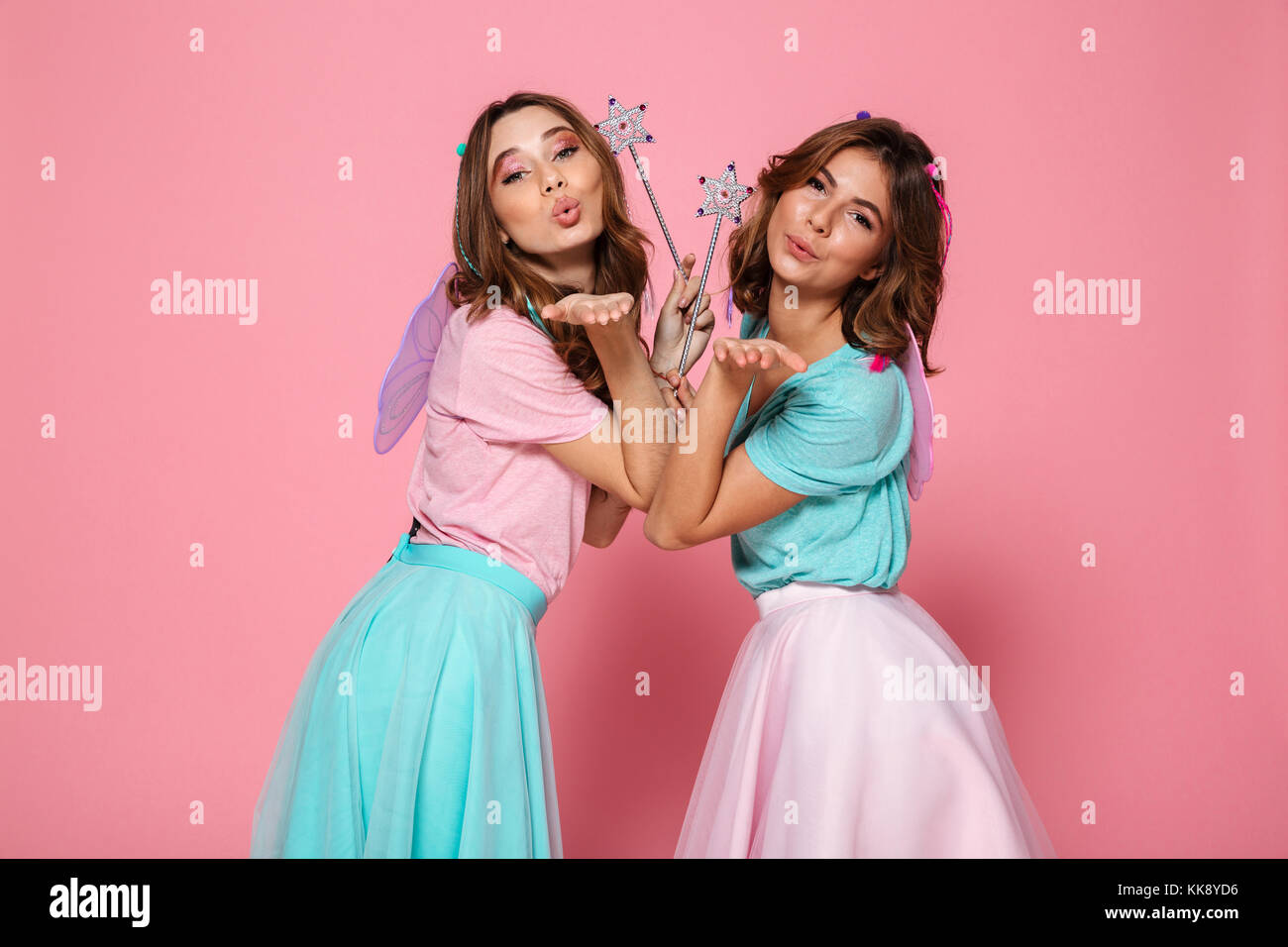 Deux jolies filles habillées comme des fées avec des ailes holding baguettes magiques tout en regardant par-dessus son épaule et le soufflage de l'appareil photo de kiss pink background Banque D'Images