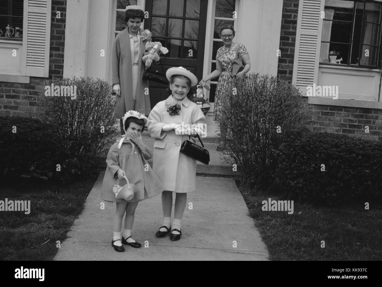 Grand-mère, fille et deux petites-filles habillés en le voyant lumineux bleu et jaune printemps de robes, de sourire et de marcher dans le chemin avant de leur maison de banlieue, 1963. Banque D'Images