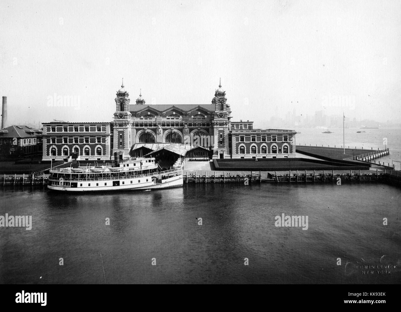 Une photographie de la façade de la station d'inspection de l'immigration d'Ellis Island, le bâtiment de style Renaissance française est construite avec des briques rouges et de calcaire de caisse, le bâtiment a ouvert ses portes le 17 décembre 1900, il a été fermé en 1954, après le traitement de plus de 12 millions d'immigrants qui entrent aux États-Unis, les tours peuvent être vus dans les coins de la partie centrale de la structure avec un passage couvert menant à l'entrée principale, un grand bateau est amarré à la fin de l'allée, New York, 1907. À partir de la Bibliothèque publique de New York. Banque D'Images