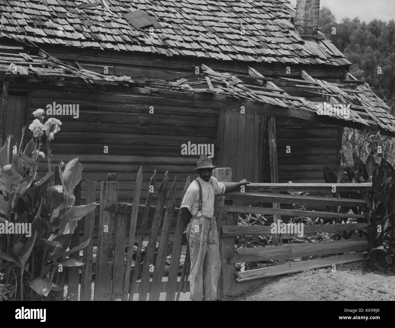 Photographie en noir et blanc d'un homme afro-américain, le port des bretelles et un chapeau, debout à une clôture en face d'une maison en bois, Août, 1940. À partir de la Bibliothèque publique de New York. Banque D'Images