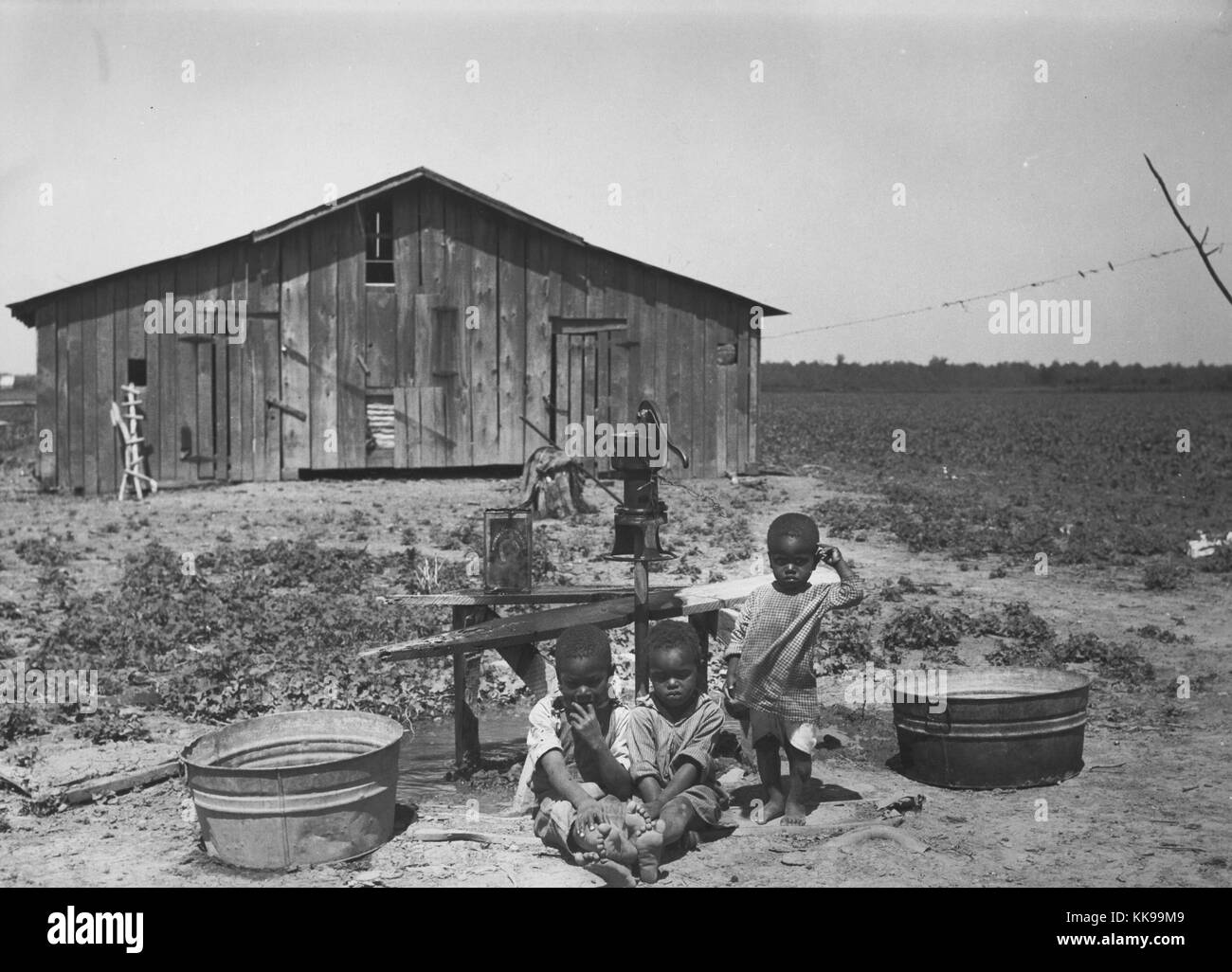 Photographie en noir et blanc de trois jeunes enfants d'origine afro-américaine, deux assis, un comité permanent, à l'extérieur, une pompe à eau et la structure en bois peut être vu dans l'arrière-plan, West Memphis, Arkansas, 1935. À partir de la Bibliothèque publique de New York. Banque D'Images