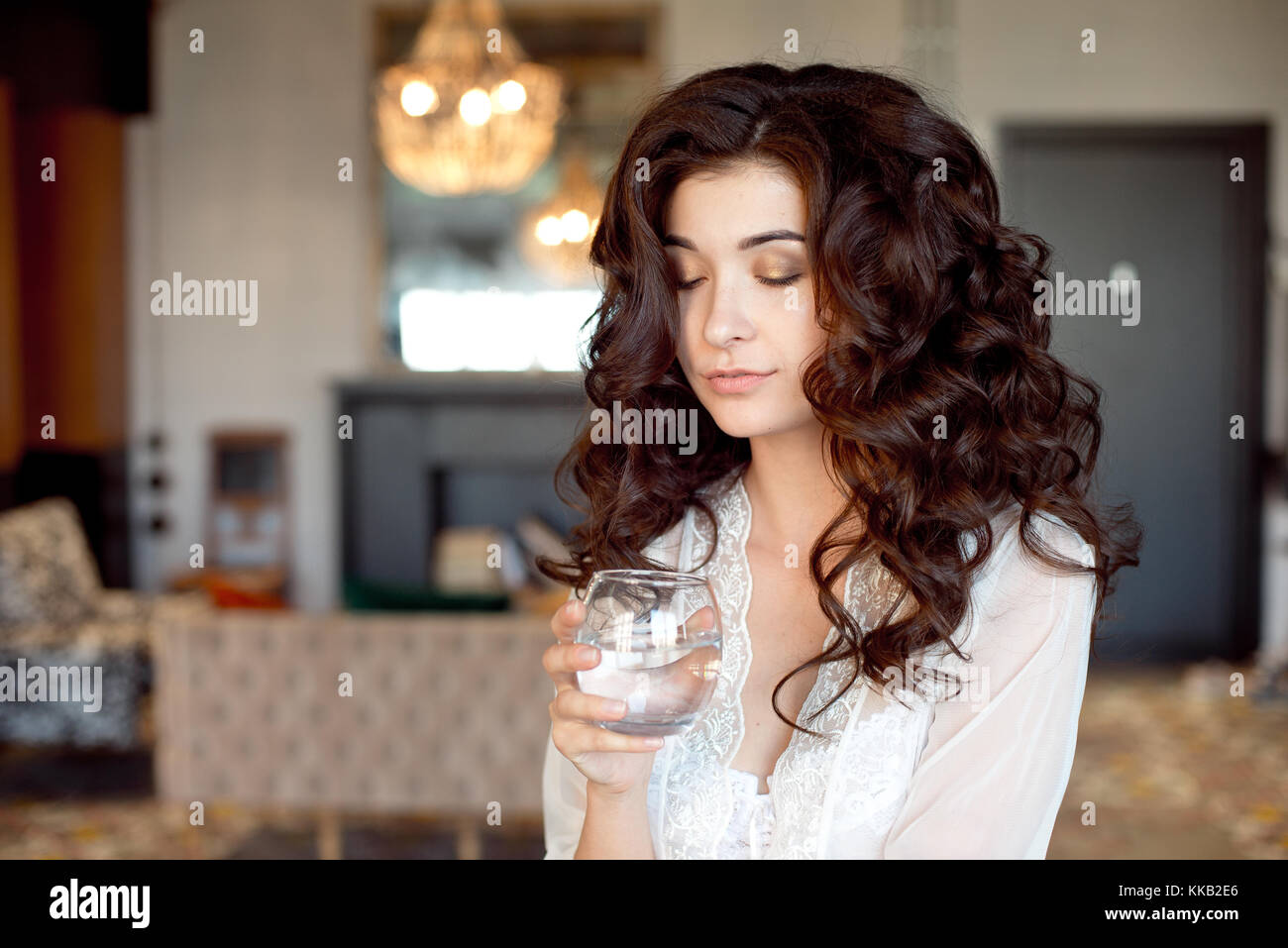 Cie jolie femme avec les yeux baissés de porter l'ombre d'oeil tenant un  verre de boisson fraîche dans sa main qu'elle siège en souriant sur un  canapé à la maison avec un